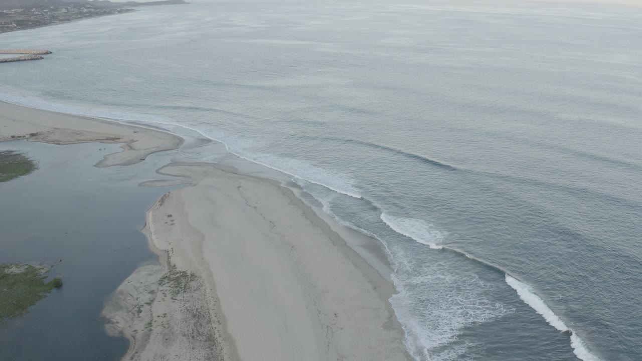 Aerial view over Los Cabos, Mexico, showing a pale sandy spit beside a dark inlet and gray-blue ocean surf, with no visible people.