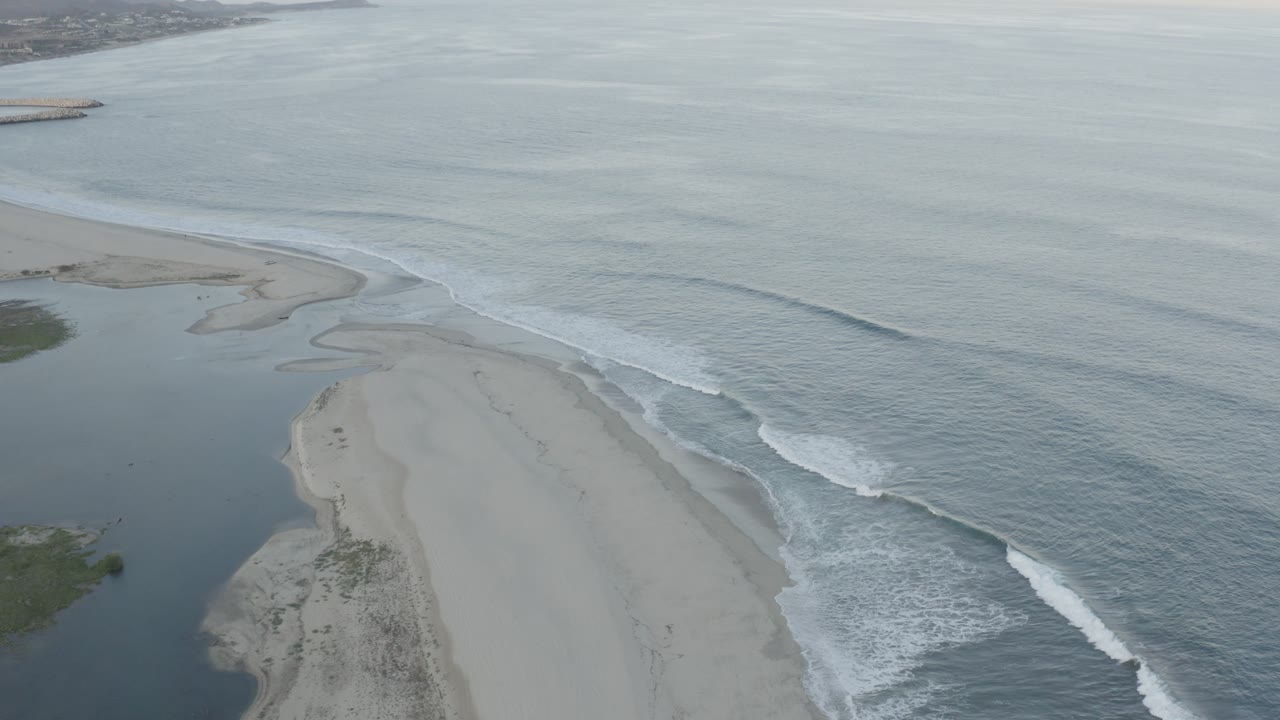 Aerial frame in Los Cabos, Mexico, with a diagonal band of white surf breaking beside the sand and a dark inlet curving along the beach, no visible people.