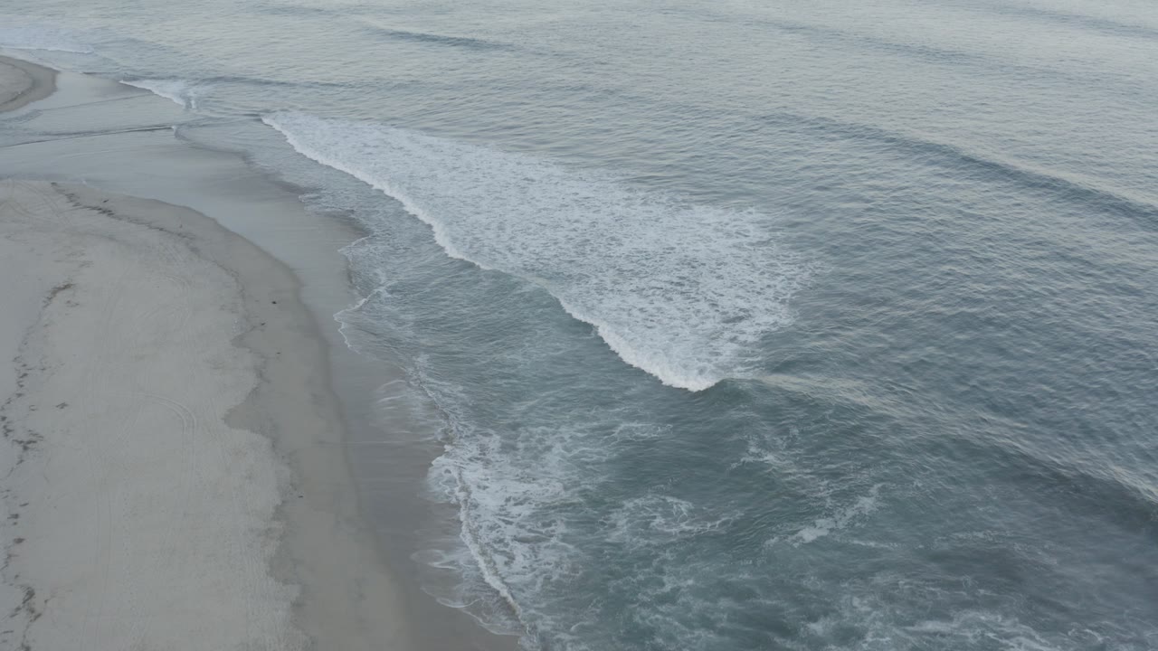 Aerial view of blue-gray surf breaking beside pale sand in Los Cabos, Mexico, with white foam angled across the shoreline and no visible people.