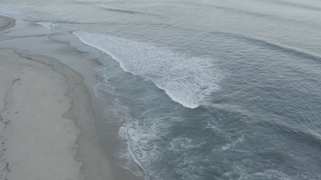 Aerial frame from Los Cabos, Mexico showing a curved white wave line pushing toward an empty sandy shore, with darker water marking the nearshore current and no visible people.