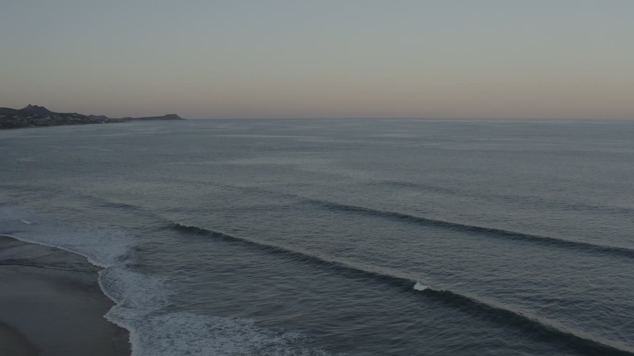 Wide elevated view of Los Cabos ocean waves angling toward a dark beach under muted early or late light, with no visible people.