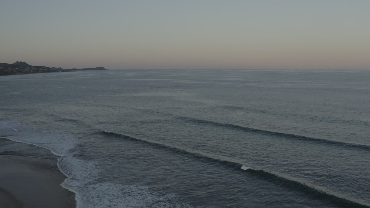 Los Cabos surf forms long parallel lines across blue-gray water, with a distant headland near the horizon and no visible people.