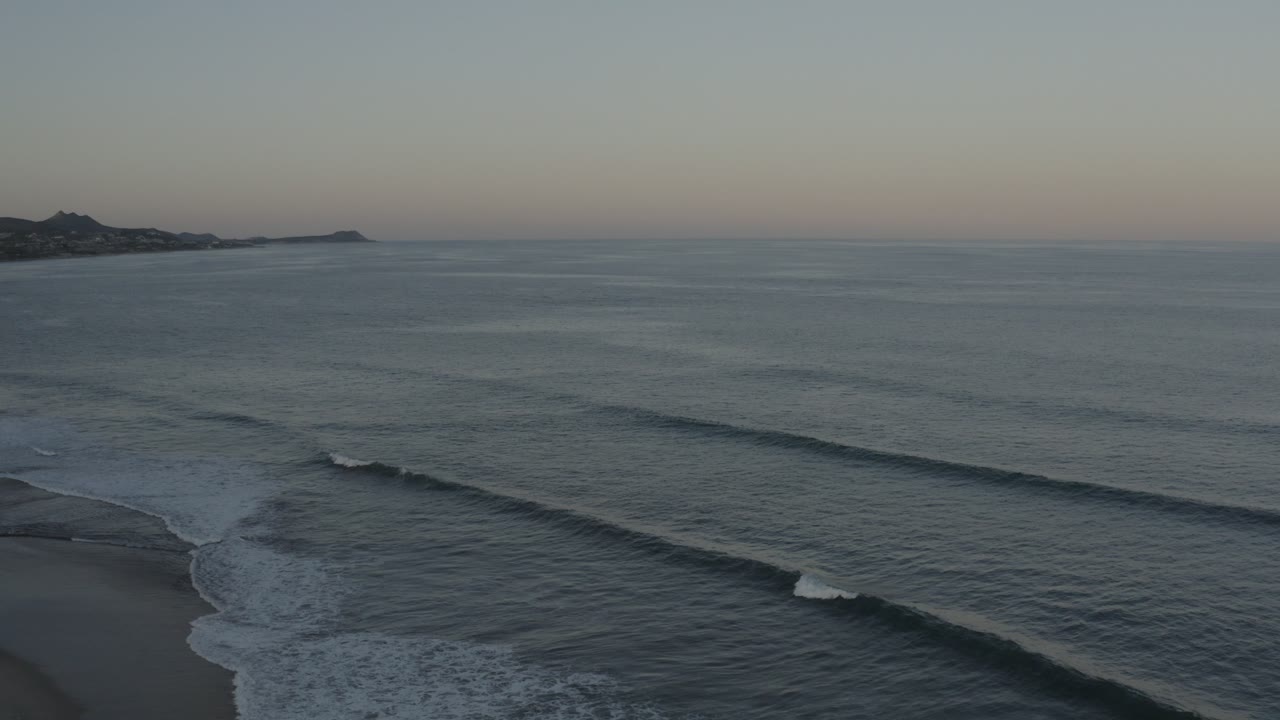 Ocean swell approaches the shoreline with low hills on the horizon.