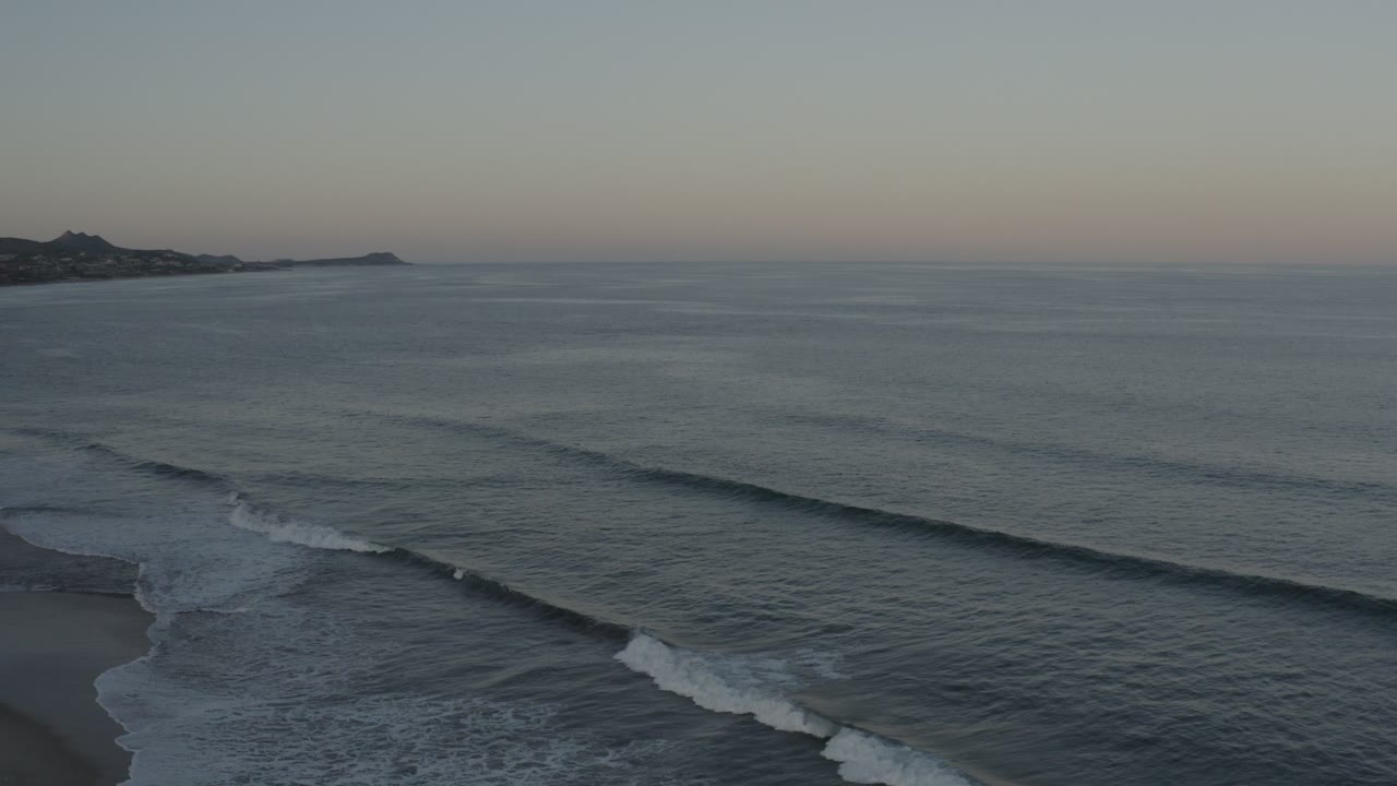 Opening aerial view in Los Cabos, Mexico, with gray-blue waves rolling toward the lower beach edge, distant dark headlands at left, soft sky color, and no visible people.