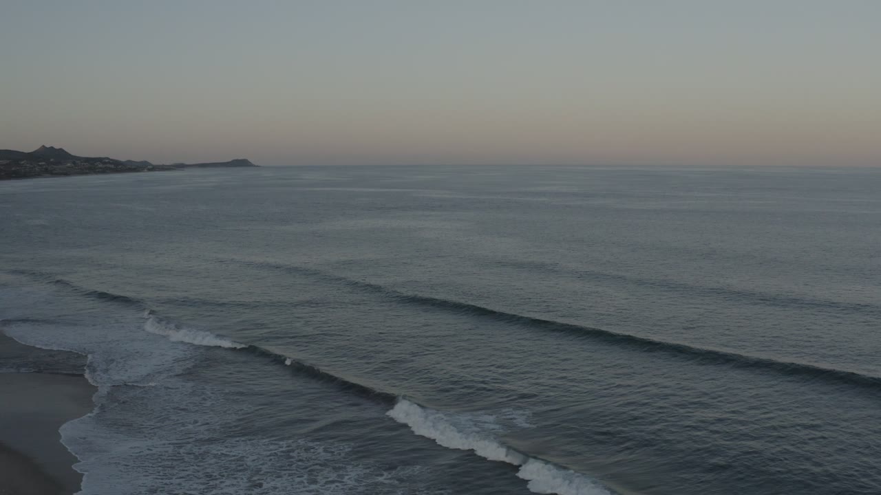 Aerial Los Cabos frame with broad gray water, parallel wave bands moving toward shore, a small dark beach area at lower left, and no visible people.