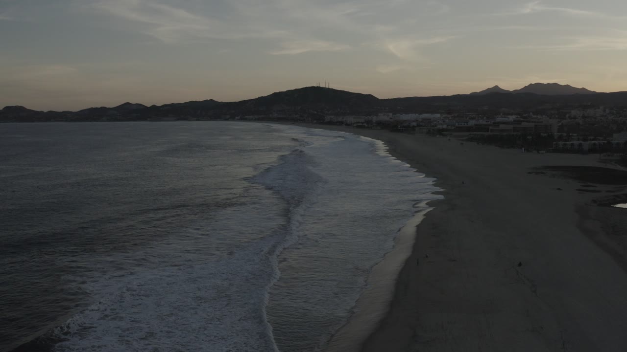 Elevated view of a dark beach shoreline with surf and mountains in the distance.