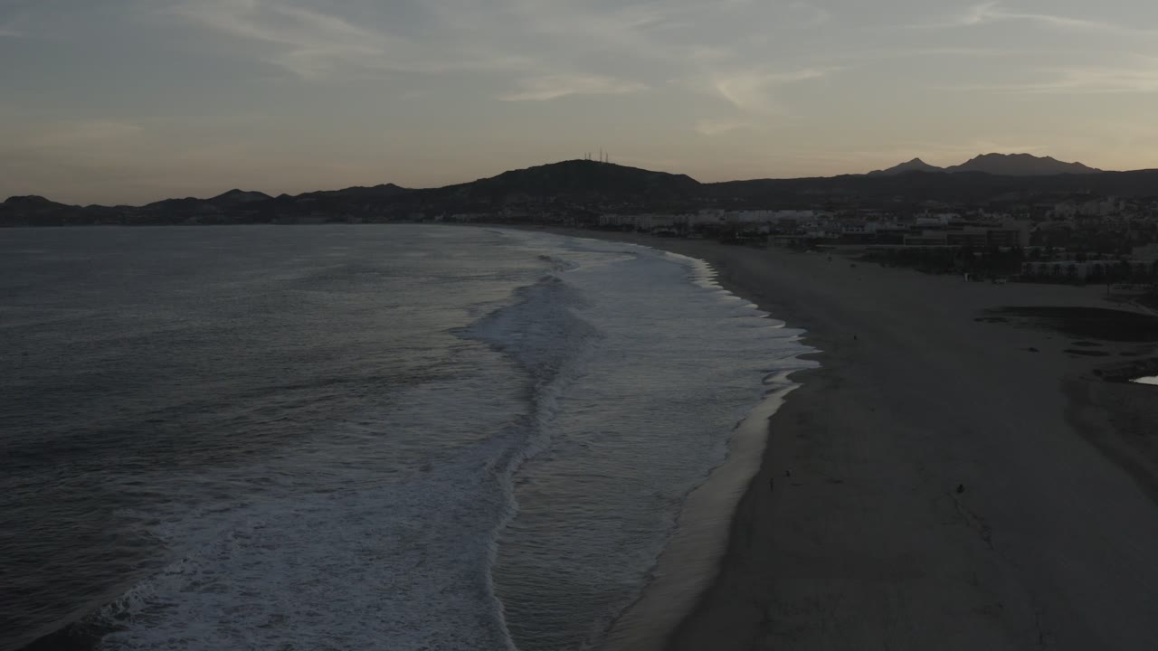 Wide Los Cabos shoreline frame with the surf band slightly farther along the beach, dark ocean texture in the foreground, distant apartments or buildings near the coast, and mountains under muted sky.