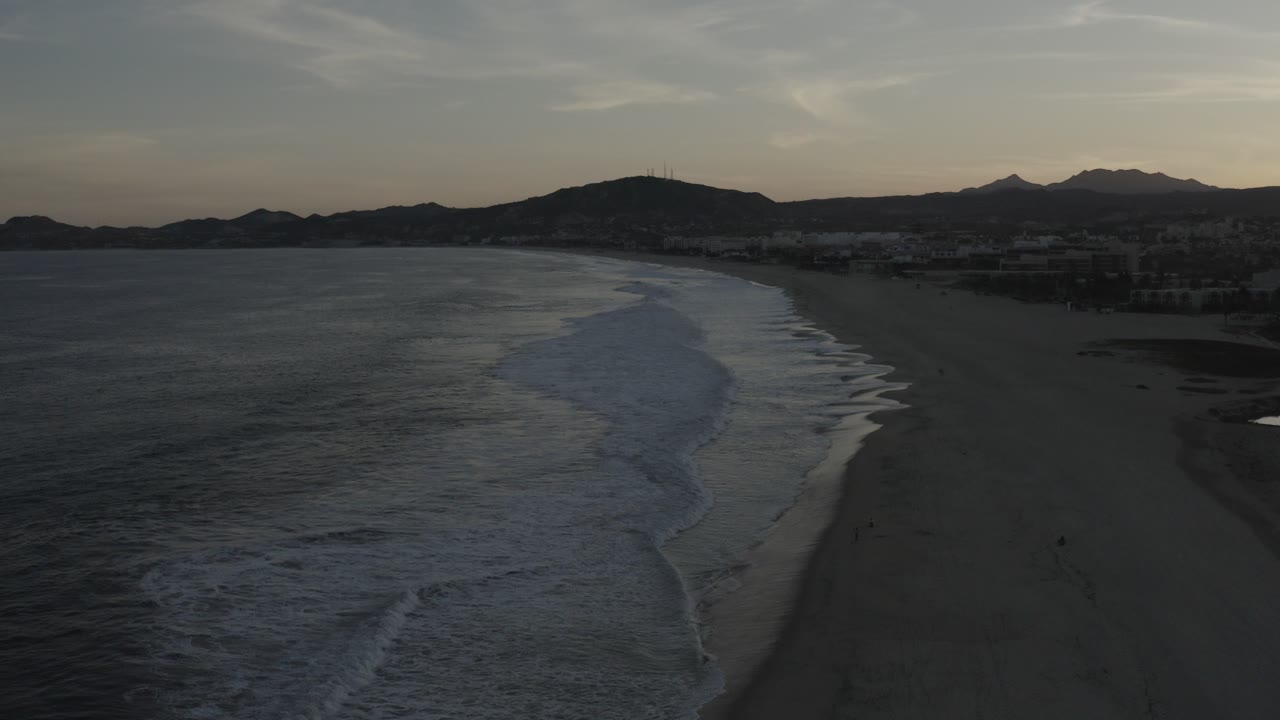 Los Cabos, Mexico aerial view with dark ocean on the left, white surf curling toward a wide beach, distant hills on the horizon, and no visible people.