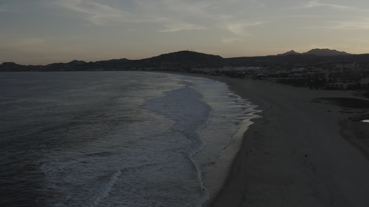 Los Cabos, Mexico coast seen from above, with a brighter diagonal band of breaking waves, dark sand to the right, hills in the background, and no visible people.