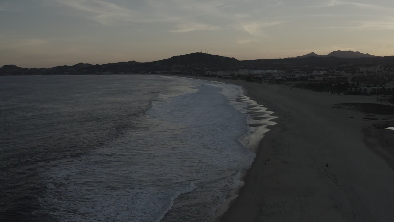 Aerial side view in Los Cabos, Mexico, with dark ocean at left, white surf bands along the beach, distant buildings, mountains, muted sky, and no visible people.