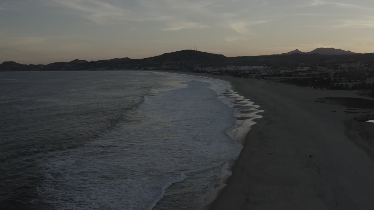 High beach view in Los Cabos, Mexico, showing the surf line closer to center frame, dark sand, distant coastal buildings, mountain silhouettes, and no visible people.
