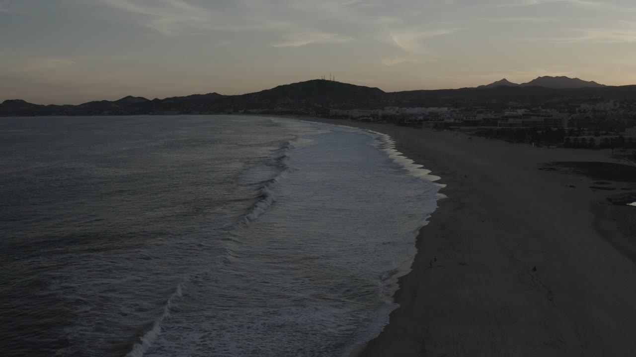 Aerial view over a Los Cabos beach at the start of the clip, with dark ocean water on the left, white surf lines on the sand, distant buildings, mountain silhouettes, and no visible people.