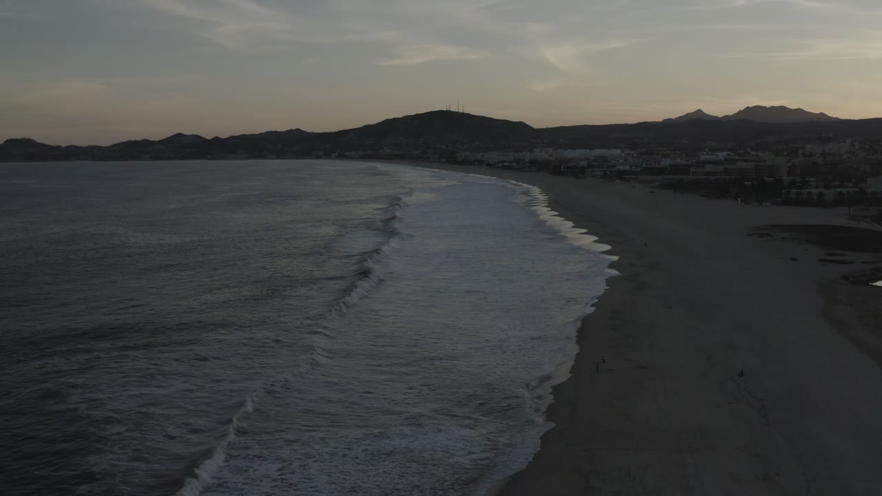 Aerial Los Cabos shoreline view one second in, with the wave break shifted farther down the beach, dark water filling the left side, sand on the right, distant coastal development, mountain silhouettes, and no visible people.