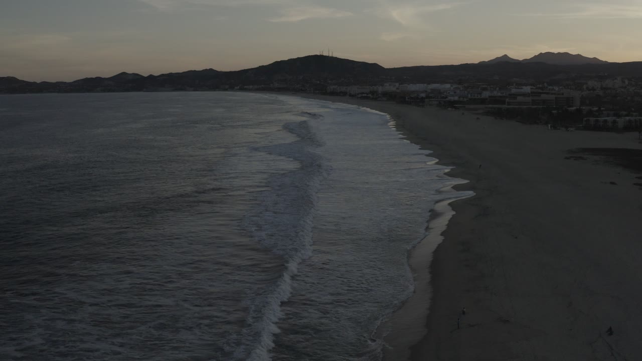 Dark dusk beach with waves and mountains in the distance.