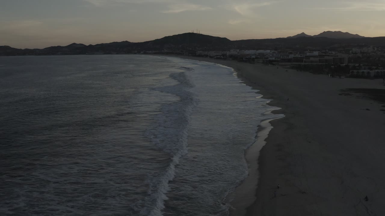 Los Cabos beach at dusk from an elevated angle, with a long band of breaking waves curving along the sand, low hills on the horizon, and no visible people.