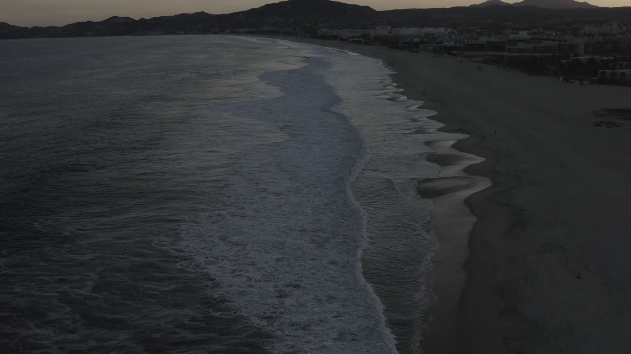 Elevated early evening view of a Los Cabos shoreline, with dark ocean water at left, foamy waves crossing the sand, distant mountains on the horizon, and no visible people.