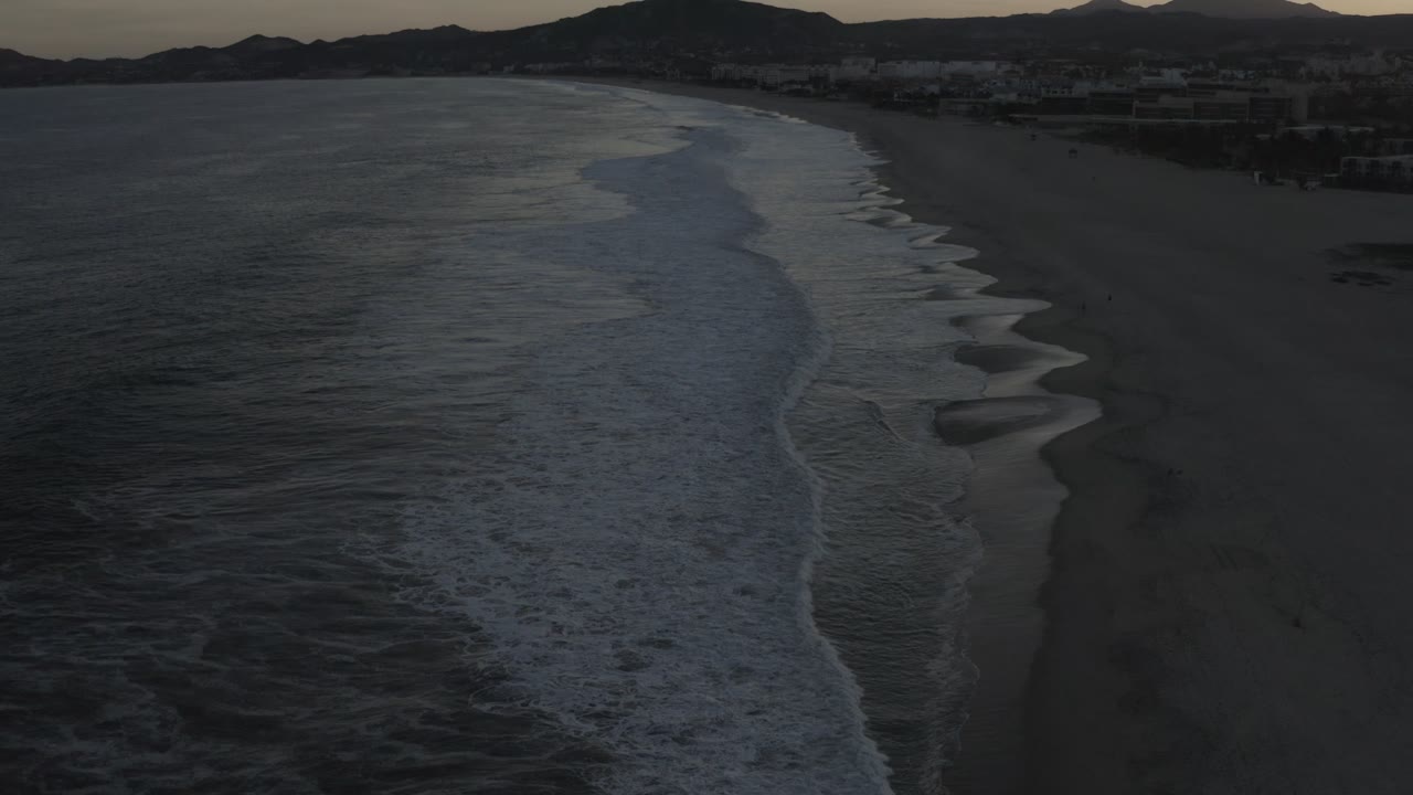 Aerial-looking view over the Los Cabos beach as white wave foam forms curved bands along the shoreline, with the sandy beach and mountain ridge fading into low light and no visible people.