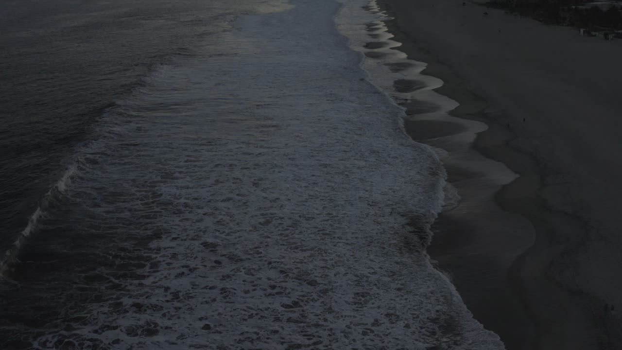 Aerial frame of broad foam bands pushing across the Los Cabos shoreline, with dark beach visible beside the water and no visible people.