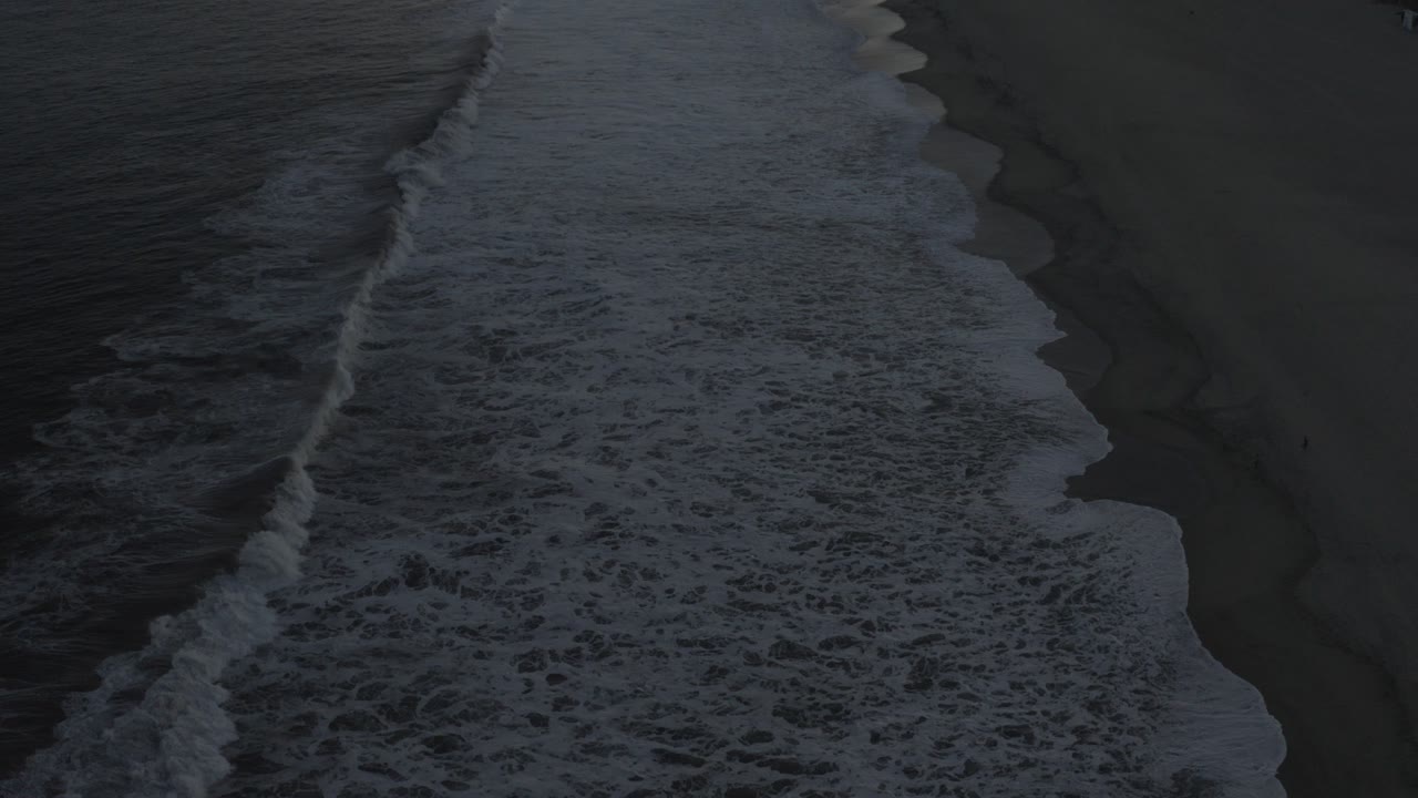 Side-top aerial view of a wide white wave breaking along a dark Los Cabos shoreline, with no visible people.