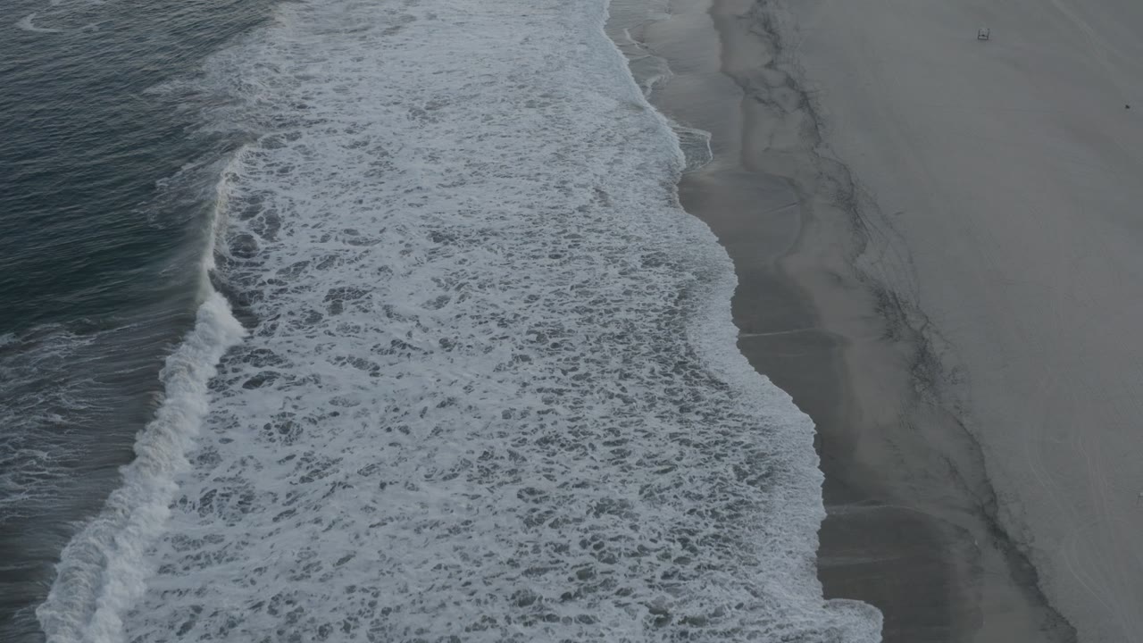 Aerial Los Cabos beach frame with a broad band of white foam pushing over wet gray sand and no visible people.