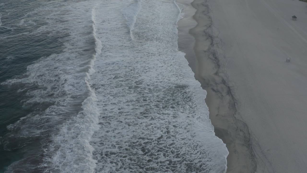 High aerial view over a Los Cabos beach, with dark water at left, white surf bands crossing the sand, and no visible people.