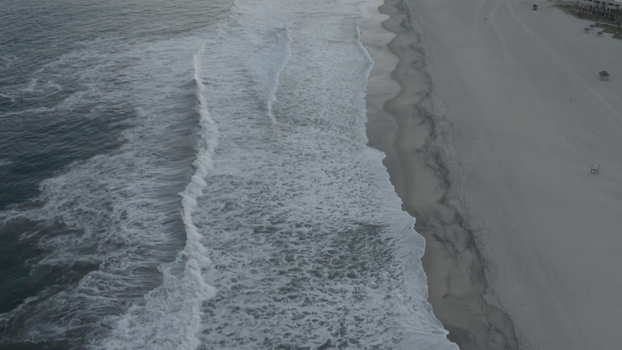 Aerial frame over Los Cabos beach with white water textured across the lower left, a narrow wet shoreline band, and broad sand beyond it, with no visible people.