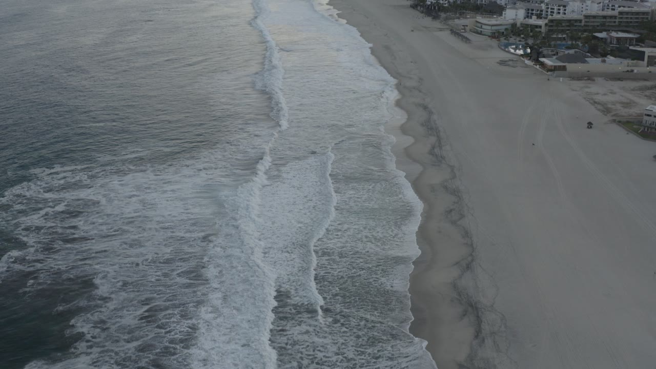 Opening aerial frame over a Los Cabos beachfront, with gray-blue ocean on the left, white surf lines, pale sand, and beachfront buildings at the upper right, with no visible people.