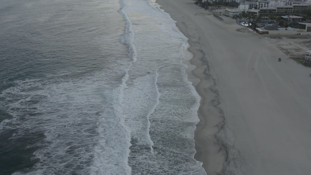Aerial view along the Los Cabos shoreline, showing the surf shifted farther down the beach, the open sand running through the center, and beachfront buildings near the top edge, with no visible people.