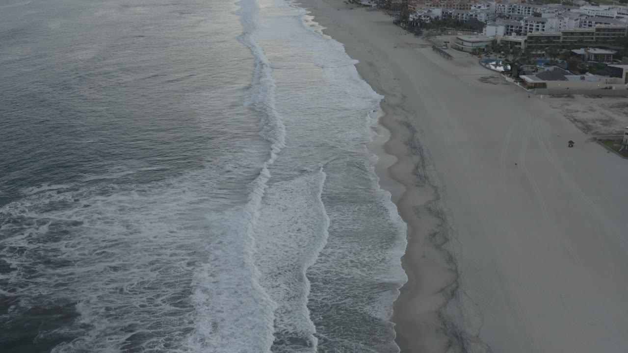 Drone view of beach surf with coastal buildings at the top of frame.