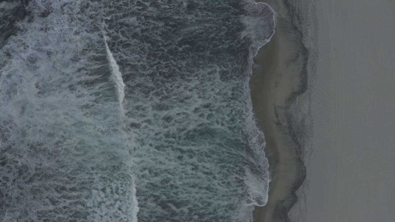 Top-down view of Los Cabos shoreline with white surf spreading over dark ocean beside wet gray sand, no visible people.