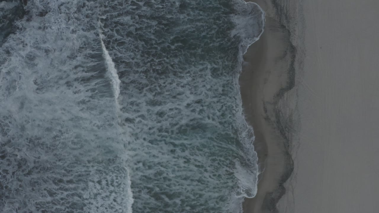 A narrow white wave crest runs through the center of the overhead Los Cabos beach view as darker water meets pale wet sand, no visible people.