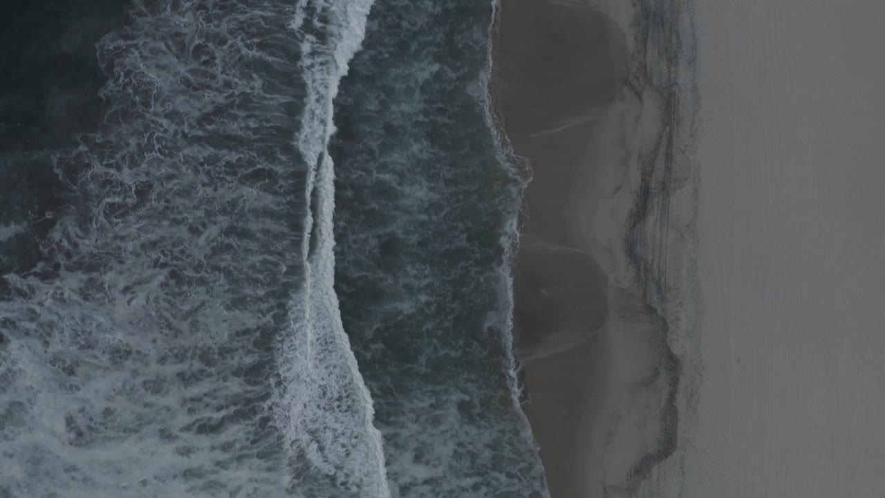 Top-down view of Los Cabos shoreline with dark ocean, white breaking foam, wet sand bands, and no visible people.