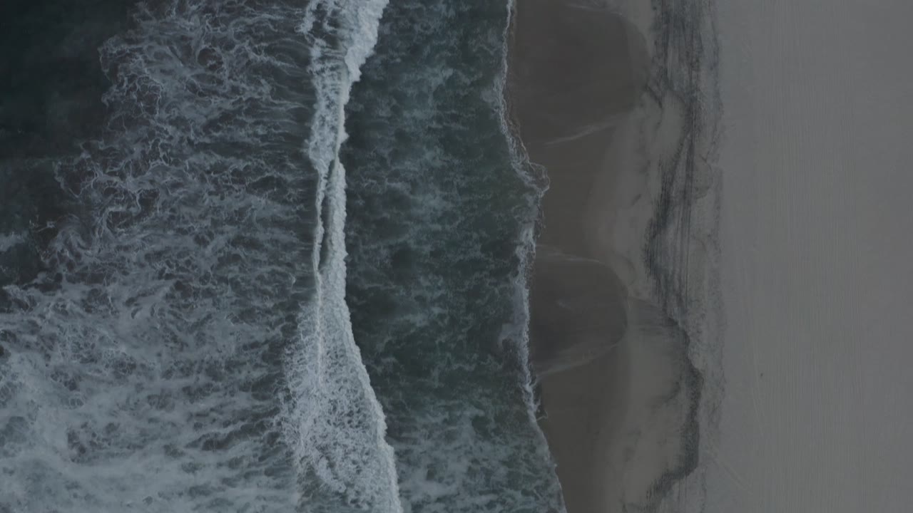Aerial Los Cabos beach frame with a wide strip of white foam over dark water and smooth sand to the side, with no visible people.