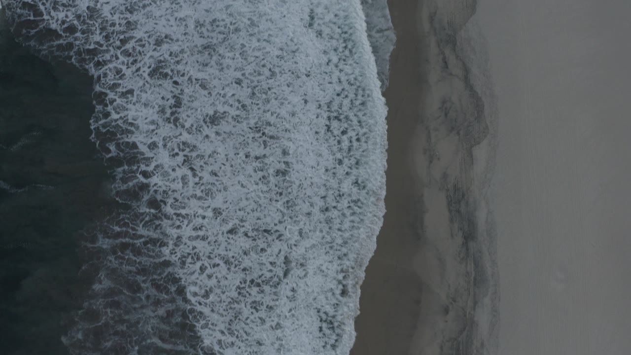 Overhead view of foamy surf meeting gray sand in Los Cabos, Mexico, with no visible people.