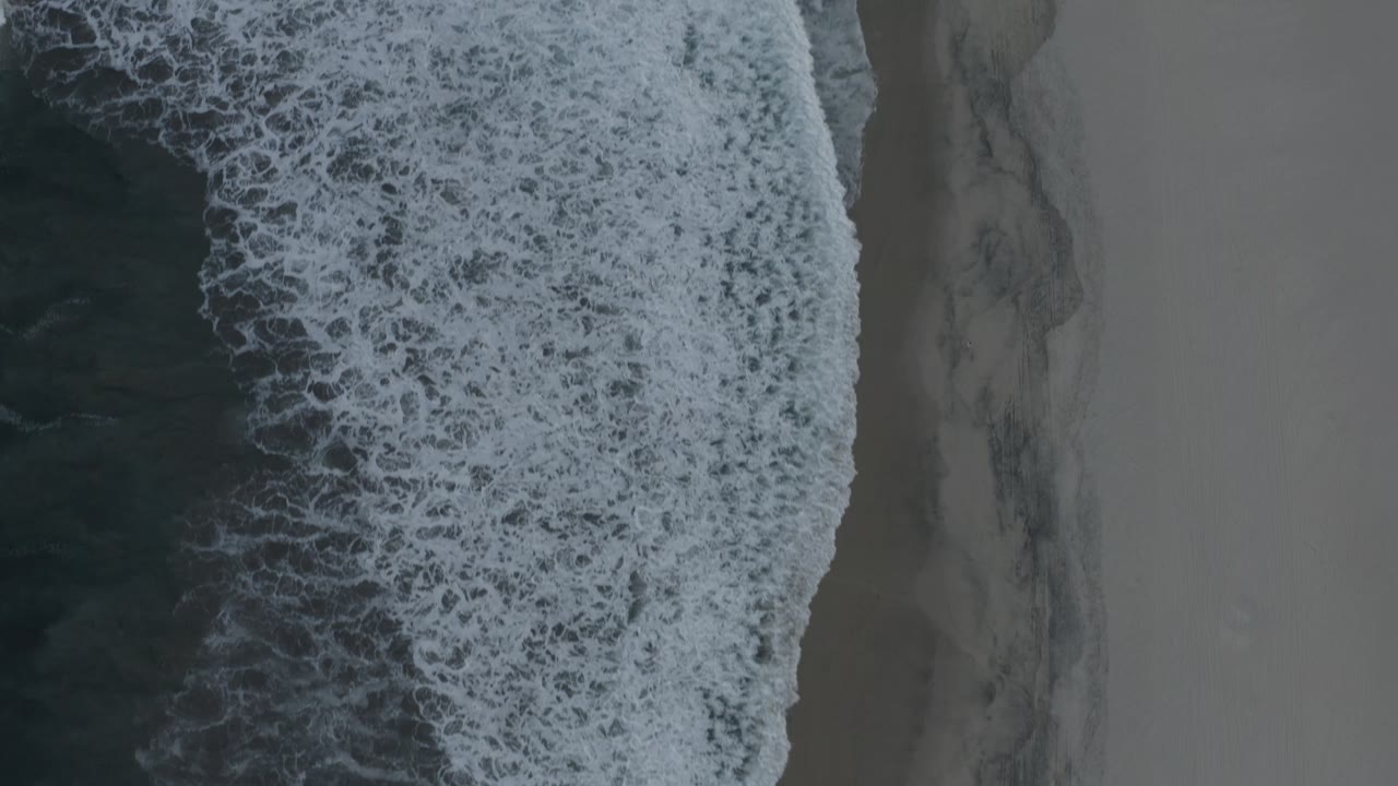 A top-down Los Cabos shoreline frame shows white foam patterns over dark water beside a narrow strip of wet sand, with no visible people.