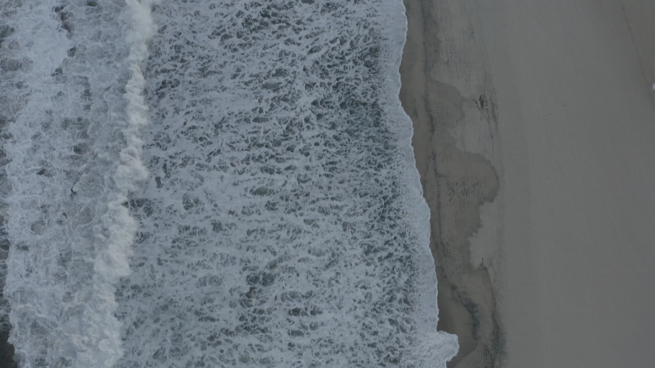Overhead view of foamy surf meeting gray sand in Los Cabos, Mexico, with no visible people at the start of the clip.