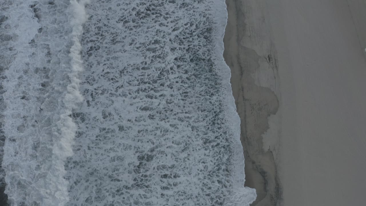 White caps spread farther across the left side of the overhead shoreline view in Los Cabos, Mexico, with no visible people.