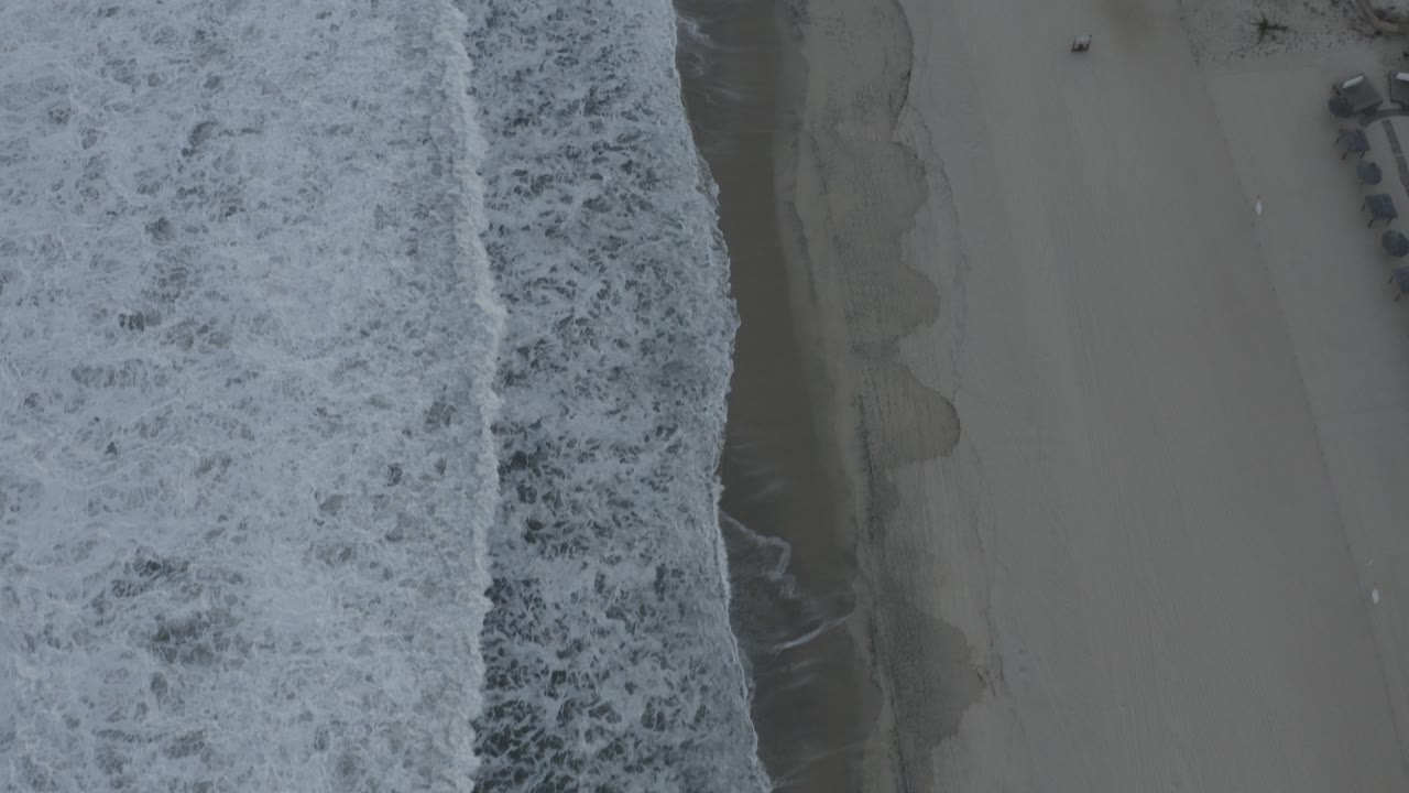 Top-down view of white surf running along gray-tan beach sand in Los Cabos, Mexico, with no visible people in the opening frame.
