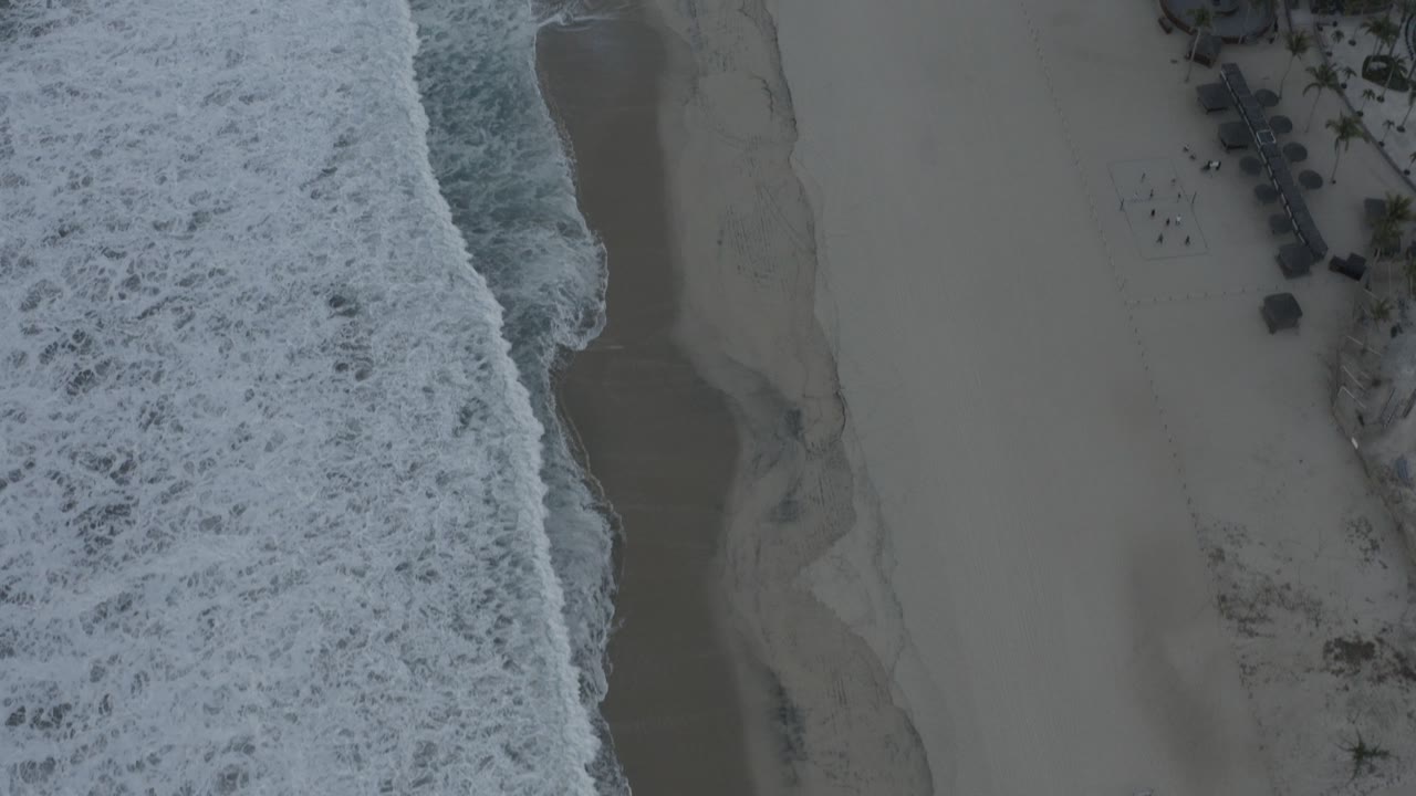 Top-down surf washing along a sandy beach beside beachfront buildings.