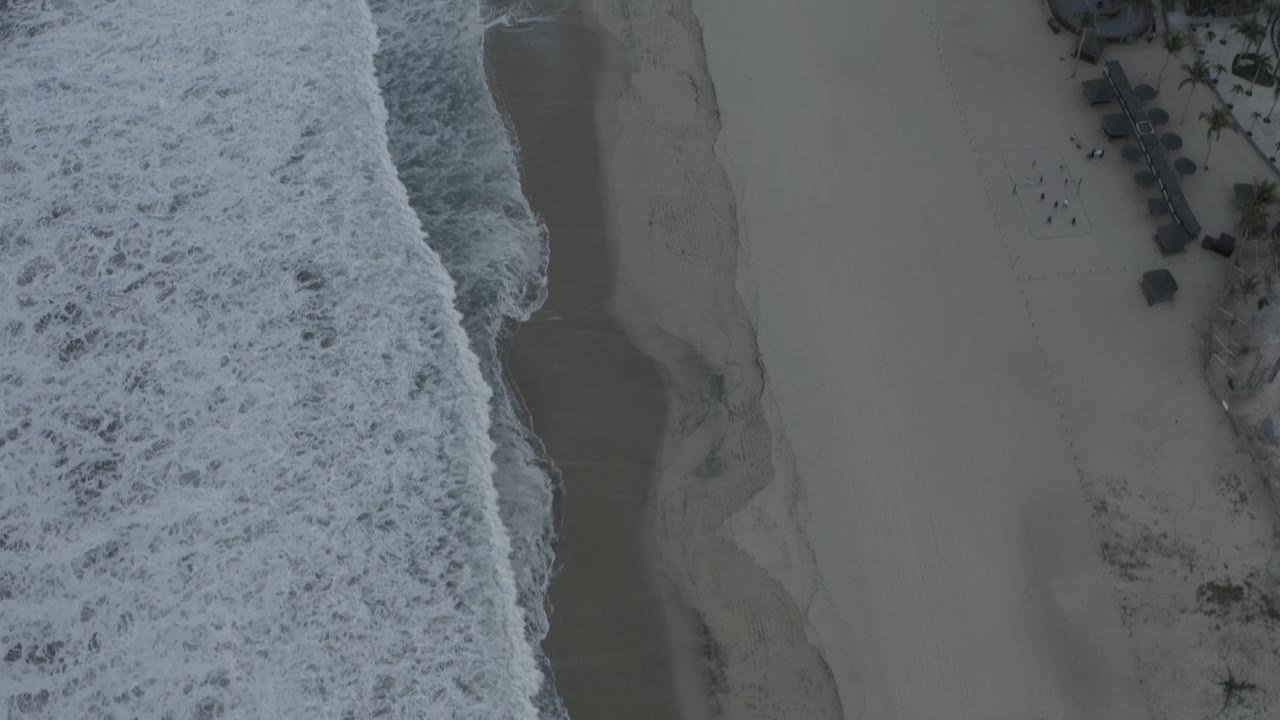 Top-down Los Cabos shoreline frame showing a wider band of foamy water, wet sand curves, small beachfront structures near the upper edge, and no visible people.
