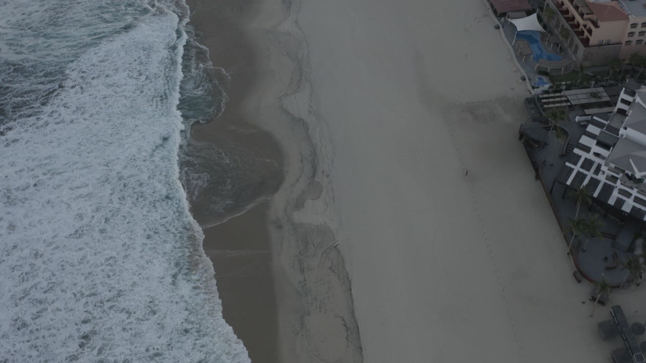 Bird's-eye view in Los Cabos, Mexico, with white surf on the left, open sand in the center, beachfront structures at the upper right, and no visible people.