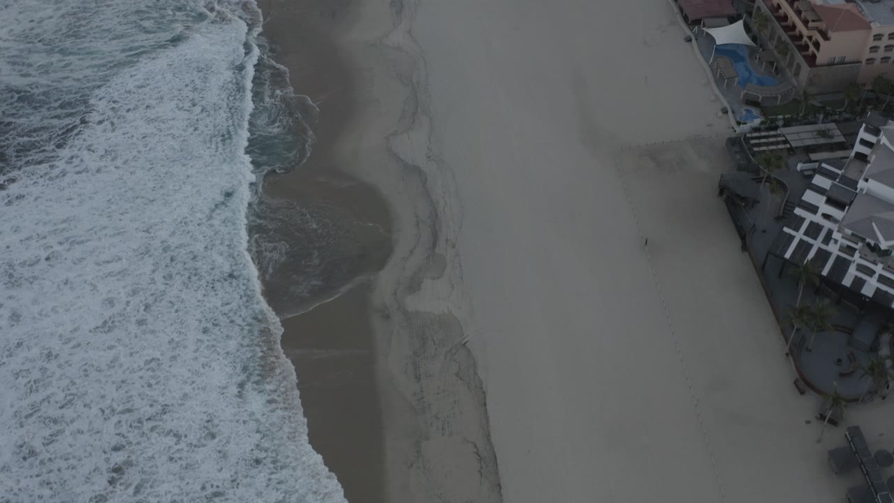 Overhead Los Cabos beach frame with the wave line closer to the center, pale sand beside it, resort-style buildings and blue pool areas near the top edge, and no visible people.