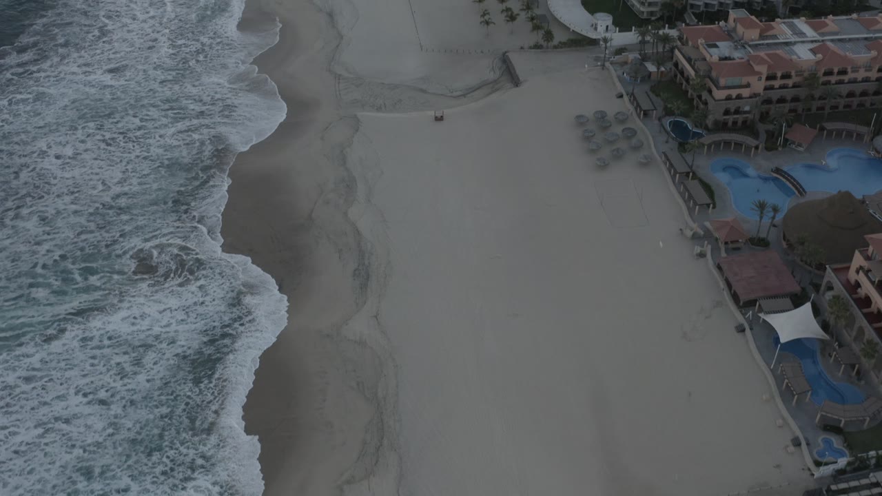 Aerial view in Los Cabos, Mexico, with the breaking shoreline running along the left edge and condominium pools and red-roofed buildings visible to the right, with no visible people.