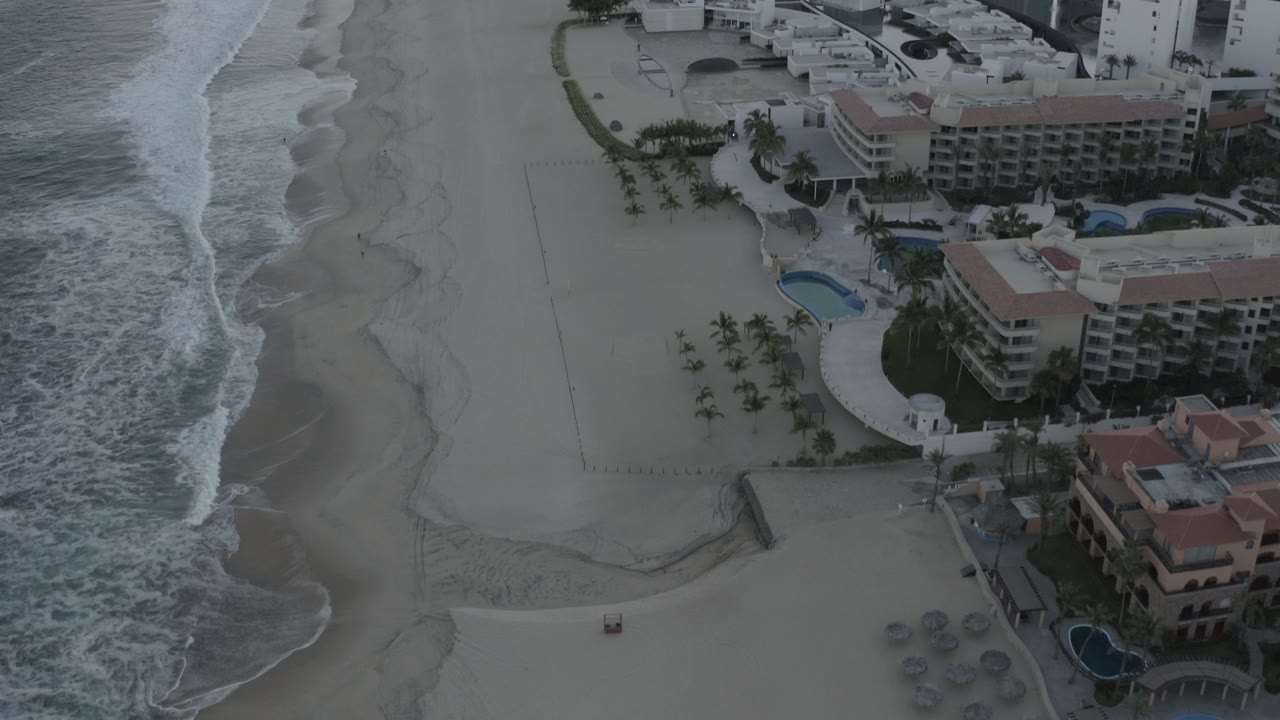 Early aerial frame over Los Cabos, Mexico showing ocean foam at left, a wide sandy beach, beachfront hotel buildings, palms, and pools with no visible people.