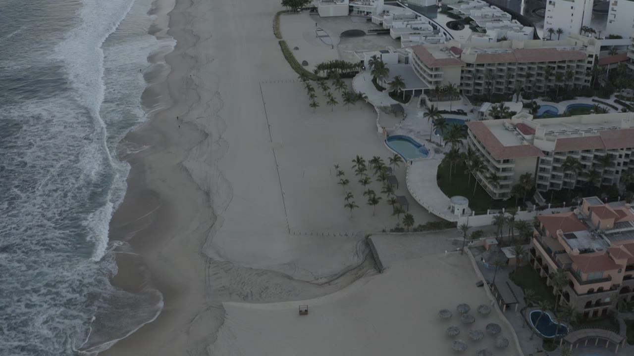 Second frame over Los Cabos, Mexico with the surf line tighter along the left edge and the beachfront hotels, blue pools, palms, and pale sand filling the right side, with no visible people.