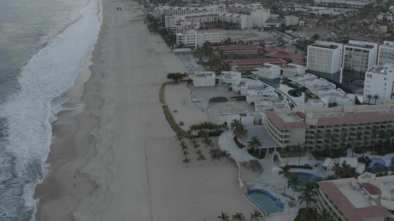 Wide aerial frame over a Los Cabos beach with surf on the left, pale sand through the center, beachfront hotels and pools at the lower right, and no visible people.