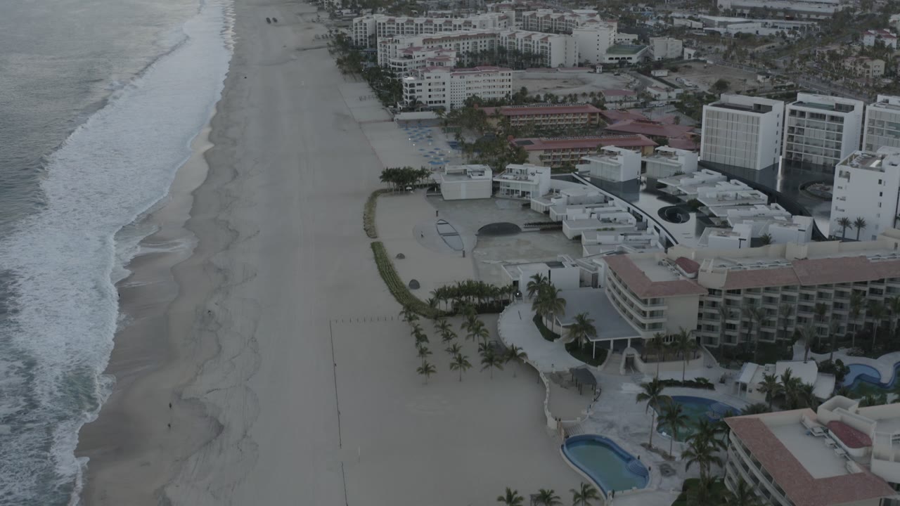 Elevated Los Cabos coastline view with white waves closer to the middle of frame, a long empty sand strip, beachfront buildings, pools, palms, and no visible people.