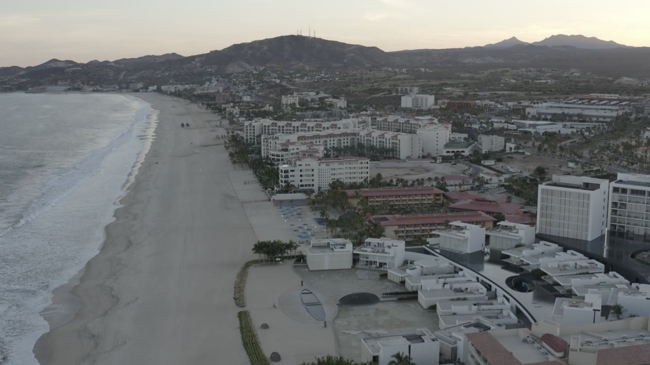 Aerial view of surf, beach, coastal buildings, and mountains.
