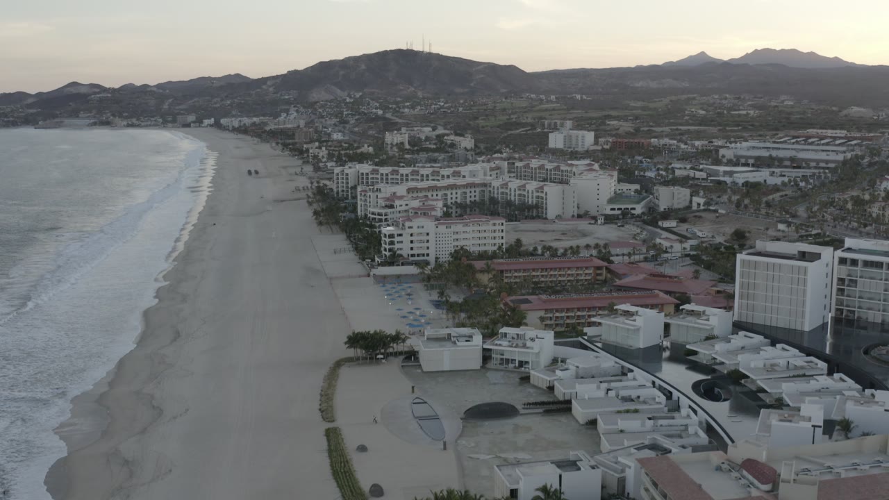 High aerial view in Los Cabos showing a broad sandy beach, rolling ocean waves, clustered beachfront buildings, inland roads, distant hills, and no visible people.