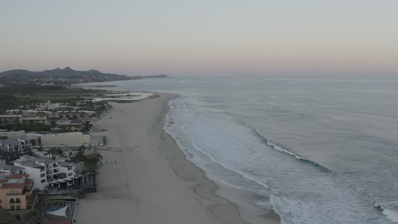 Aerial frame of the Los Cabos shoreline with the surf line slightly farther down the beach, distant hills on the horizon, beachfront structures at lower left, and no visible people.